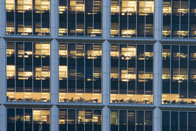 Front facade of a modern glass-walled office block at night with lights on in the offices showing staff working late into the evening